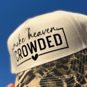 May include: A beige baseball cap with the text "make heaven CROWDED" in black, with a black heart symbol. The brim features a camouflage leopard print design. The background is a clear blue sky.