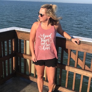May include: A woman wearing a pink tank top with the text "Good Times and Tan Lines" and a black lace-trimmed shorts stands on a wooden deck overlooking the ocean.