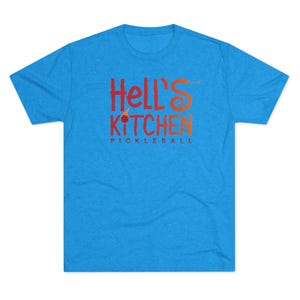 May include: A bright blue short-sleeved t-shirt featuring the red and orange text "Hell's Kitchen Pickleball" printed on the chest. The letter S in Hell's is stylized as a snake, and the letter I in Kitchen includes a small pickleball graphic.