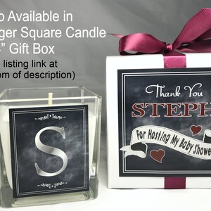 May include: A square glass candle jar with a white candle and a black and silver label with the letter "S". The candle is in a white gift box with a burgundy ribbon and a black and white label that says "Thank You STEPH For Hosting My Baby Shower".