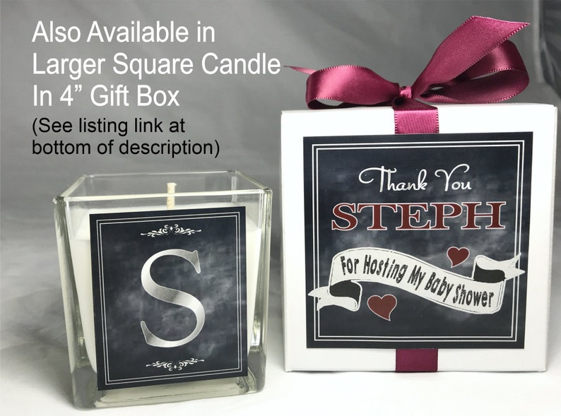 May include: A square glass candle jar with a white candle and a black and silver label with the letter "S". The candle is in a white gift box with a burgundy ribbon and a black and white label that says "Thank You STEPH For Hosting My Baby Shower".
