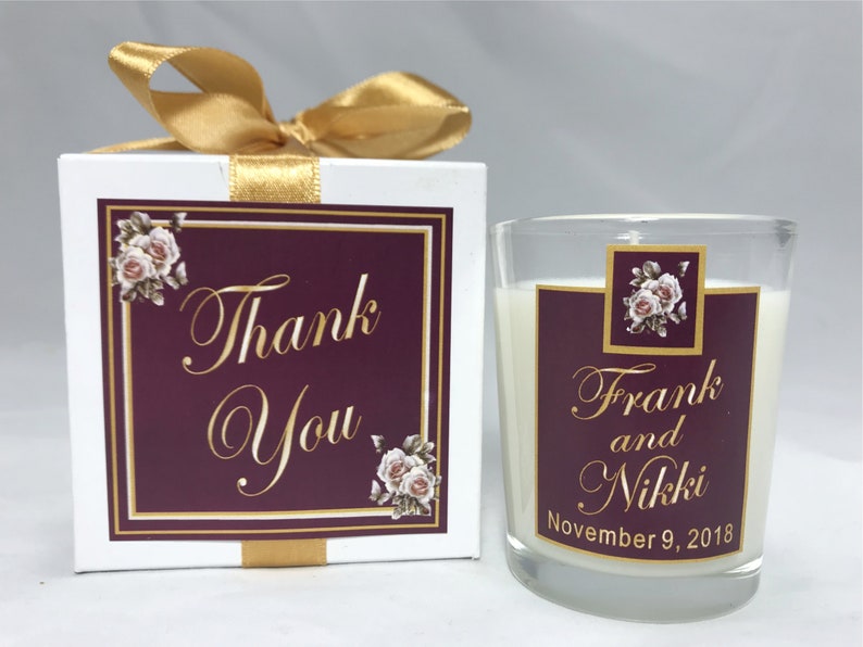May include: A white box with a gold ribbon and a burgundy label that says "Thank You" in gold lettering. The box contains a small glass jar candle with a burgundy label that says "Frank and Nikki November 9, 2018" in gold lettering.