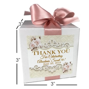 Op de afbeelding: Witte bedankbox met een roze satijnen lint en strik. De doos heeft een decoratief label met een bloemenontwerp en de tekst "THANK YOU For Celebrating Alexdents' Sweet 16! June 25, 2019". De doos meet 7,6 cm bij 7,6 cm bij 7,6 cm.