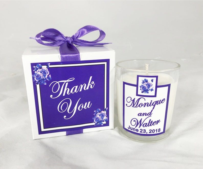 May include: A small white candle in a clear glass jar with a purple and white label that says "Monique and Walter June 23, 2018". The candle is in a white box with a purple ribbon and a purple and white label that says "Thank You".