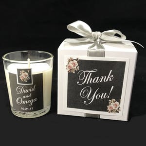 May include: A white candle in a glass jar with a black label that says "David and Omega 10.21.17". The candle is in a white box with a silver ribbon and a black label that says "Thank You!" with a floral design.