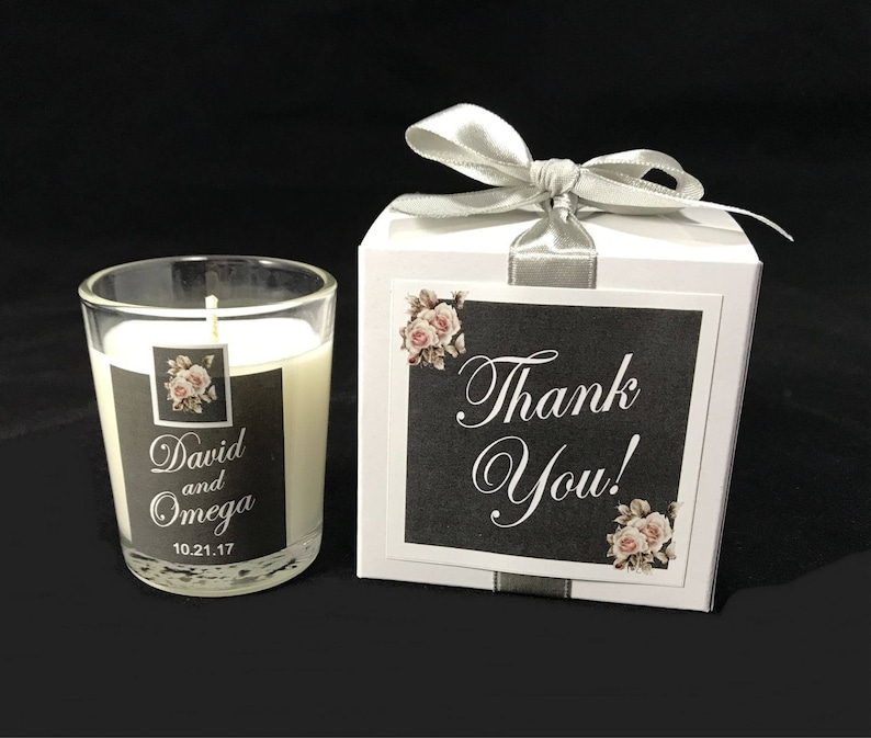 May include: A white candle in a glass jar with a black label that says "David and Omega 10.21.17". The candle is in a white box with a silver ribbon and a black label that says "Thank You!" with a floral design.