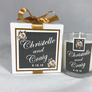 May include: A white candle in a clear glass jar with a gray and white label that says "Christelle and Craig 8.18.18". The candle is in a white box with a gold ribbon and a similar label.