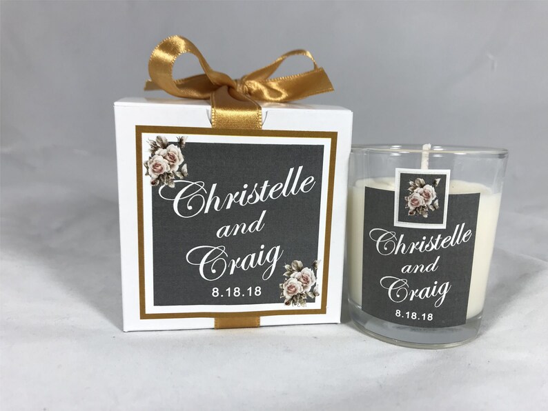 May include: A white candle in a clear glass jar with a gray and white label that says "Christelle and Craig 8.18.18". The candle is in a white box with a gold ribbon and a similar label.