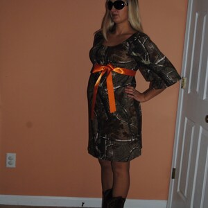 May include: A woman wearing a camouflage print dress with an orange ribbon tied around the waist. She is wearing black sunglasses and cowboy boots.