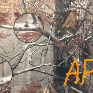 May include: Camouflage fabric with the text " Realtree" and "30" printed on it. The fabric has a brown, green, and tan color scheme. The letters "AP" are written in orange on the fabric.