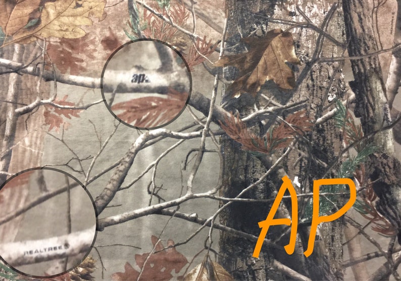 May include: Camouflage fabric with the text " Realtree" and "30" printed on it. The fabric has a brown, green, and tan color scheme. The letters "AP" are written in orange on the fabric.