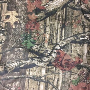 May include: Camouflage fabric with a brown, green, and red pattern. The pattern features branches, leaves, and tree trunks.