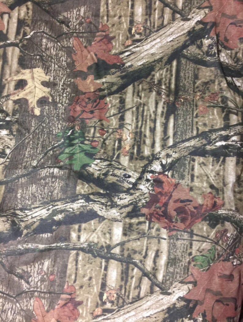 May include: Camouflage fabric with a brown, green, and red pattern. The pattern features branches, leaves, and tree trunks.