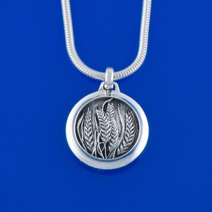 May include: Silver-toned pendant necklace with a wheat design. The round pendant features a detailed relief of wheat stalks. The necklace has a snake chain and is set against a blue background.