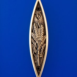 May include: An elongated, gold-toned metal decorative piece with a wheat stalk design. The wheat is detailed with a dark, textured finish, set against a bright blue background. The piece has a pointed top and bottom.