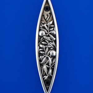 May include: A silver-toned, elongated decorative piece with a botanical design. The piece features a central carved pattern of leaves, berries, and a Hebrew letter, with black square accents at each end. The background is a vibrant blue.