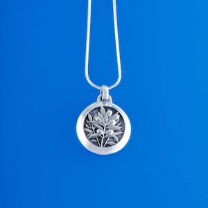May include: Silver-toned pendant necklace featuring a circular pendant with a detailed olive branch design. The pendant hangs from a delicate silver chain against a vibrant blue background. A classic piece of jewelry.