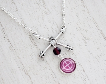 Puede incluir: Collar plateado con flechas cruzadas, una pequeña cuenta roja oscura y un colgante circular con un diseño rosa. La cadena es delicada, y el colgante tiene un diseño único y simbólico.