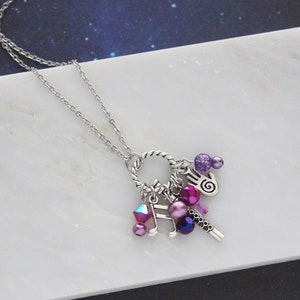 May include: A silver chain necklace with a charm bracelet featuring a variety of charms, including a music note, a hand, and several purple, pink, and blue beads.