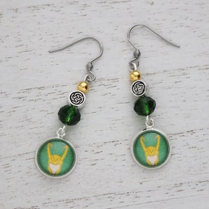 以下が含まれることがあります： 緑と金のアクセントが施された、シルバーカラーのぶら下がりピアス。各ピアスには、スタイリッシュなデザインの緑と金の円形チャーム、緑色のビーズ、ケルトノットチャームが付いています。