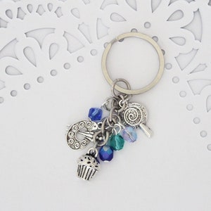 May include: A silver key chain with a cupcake charm, a paintbrush charm, a lollipop charm, and blue and green crystal beads.
