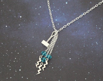 Puede incluir: Un collar de plata con un dije de martillo de plata y cuentas de cristal azul que cuelgan de él. El collar está sobre un fondo azul oscuro con estrellas blancas.