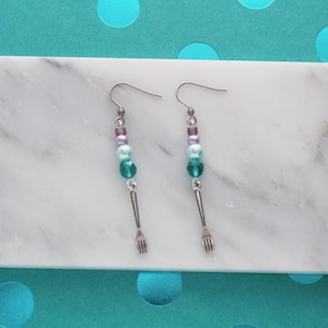May include: A pair of dangle earrings with a culinary theme. Each earring features a silver-colored fork charm suspended below a strand of beads in shades of teal, purple, and light blue. The earrings are displayed on a white marble surface.
