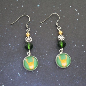 以下が含まれることがあります： シルバーカラーのドロップピアス。各イヤリングには、手のジェスチャーデザインの緑と金の円形チャームが付いています。イヤリングには、緑と金のビーズ、ケルトノットチャームも含まれています。