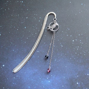 Puede incluir: Marcap&aacute;ginas de plata con un colgante de un p&aacute;jaro con cuentas de cristal negras y rojas.