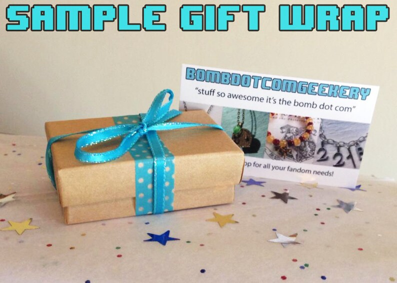 以下が含まれることがあります： 小さな茶色のギフトボックスで、白い水玉模様のターコイズリボンで結ばれています。ボックスは、カラフルな星型の紙吹雪のある表面にあります。「SAMPLE GIFT WRAP」と「BOMBOOTCOMGEEKERY」というテキストのカードが背景にあります。