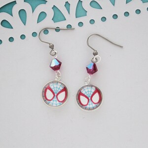 Puede incluir: Un par de pendientes colgantes plateados. Cada pendiente presenta un dise&ntilde;o de m&aacute;scara de Spider-Man en rojo, blanco y azul. Una cuenta de cristal rosa oscuro facetada cuelga sobre el dije. Los pendientes tienen un estilo de gancho de pez.