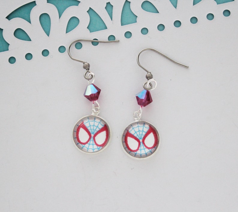 Puede incluir: Un par de pendientes colgantes plateados. Cada pendiente presenta un dise&ntilde;o de m&aacute;scara de Spider-Man en rojo, blanco y azul. Una cuenta de cristal rosa oscuro facetada cuelga sobre el dije. Los pendientes tienen un estilo de gancho de pez.