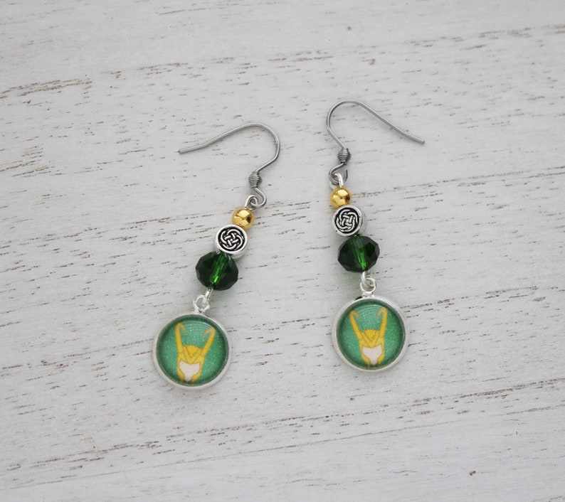以下が含まれることがあります： 緑と金のアクセントが施された、シルバーカラーのぶら下がりピアス。各ピアスには、スタイリッシュなデザインの緑と金の円形チャーム、緑色のビーズ、ケルトノットチャームが付いています。