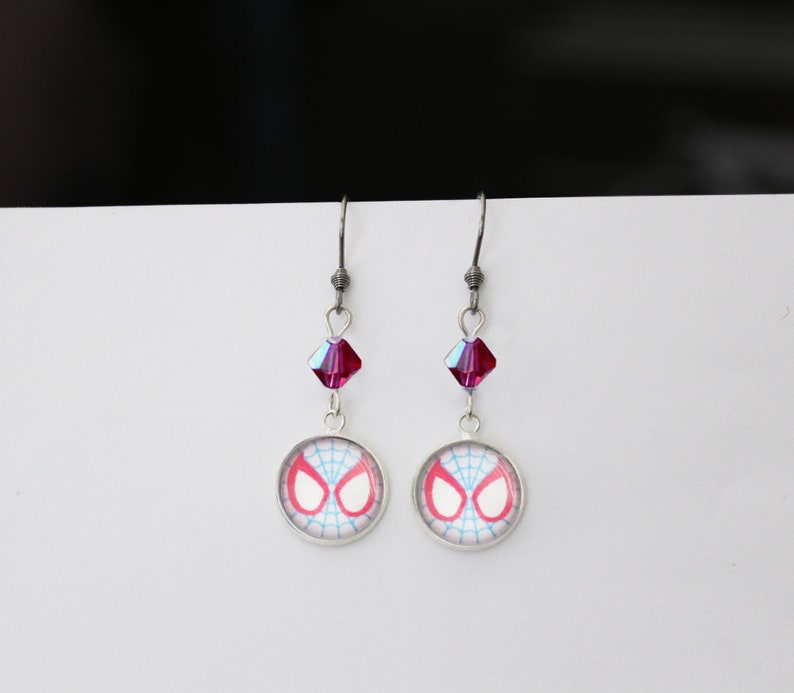 Puede incluir: Un par de pendientes colgantes plateados. Cada pendiente presenta un dije redondo con un dise&ntilde;o de ojos de ara&ntilde;a rojos y blancos, una cuenta de cristal magenta y un gancho plateado. Los pendientes se muestran sobre un fondo blanco.