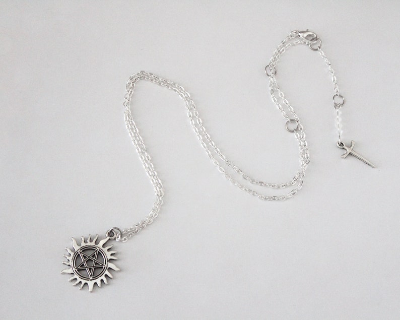 K&ouml;nnte beinhalten: Silberne Halskette mit einem Pentagramm-Anh&auml;nger und einem Schwertanh&auml;nger. Der Anh&auml;nger ist ein f&uuml;nfzackiger Stern, der von einem Kreis umgeben ist, und der Schwertanh&auml;nger ist ein kleines, schlichtes Schwert.