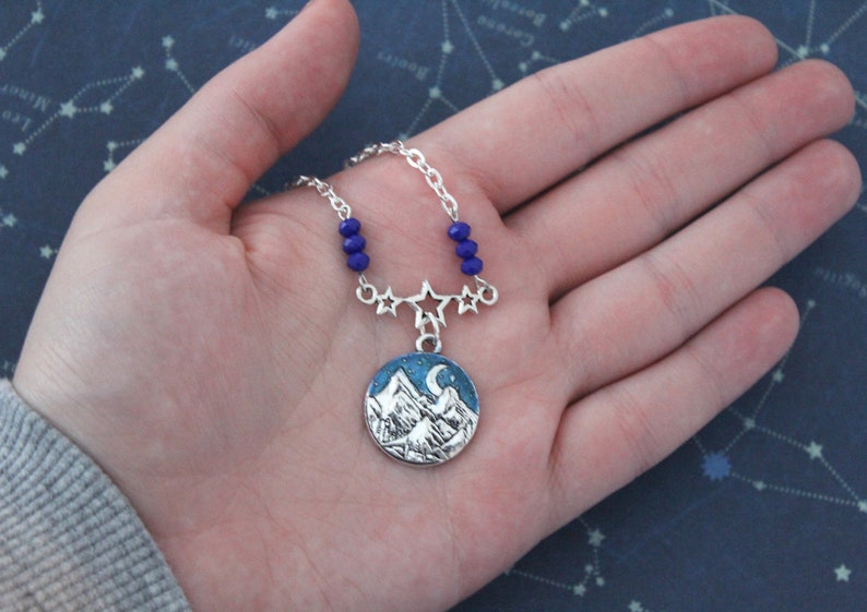 May include: A silver necklace with a blue and white mountain scene pendant. The pendant features a crescent moon and stars. The necklace is adorned with blue beads and silver star charms.