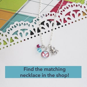 Puede incluir: Un collar plateado con tres colgantes: un colgante de Spider-Man, un charm de ara&ntilde;a y dos cuentas de colores. El collar se muestra sobre una superficie blanca con un borde de papel decorativo. Texto: "Find the matching necklace in the shop!"