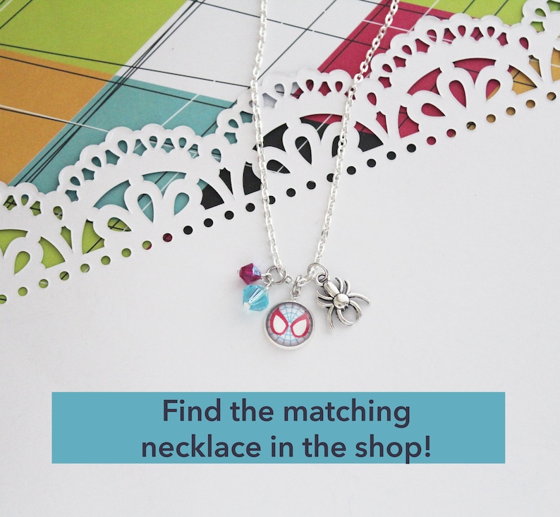 Puede incluir: Un collar plateado con tres colgantes: un colgante de Spider-Man, un charm de ara&ntilde;a y dos cuentas de colores. El collar se muestra sobre una superficie blanca con un borde de papel decorativo. Texto: "Find the matching necklace in the shop!"