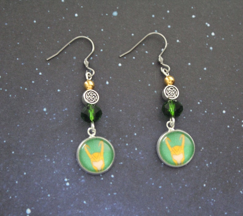 以下が含まれることがあります： シルバーカラーのドロップピアス。各イヤリングには、手のジェスチャーデザインの緑と金の円形チャームが付いています。イヤリングには、緑と金のビーズ、ケルトノットチャームも含まれています。