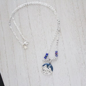 May include: A silver chain necklace with a silver charm featuring a mountain scene with a crescent moon and stars. The charm is surrounded by blue iridescent beads.