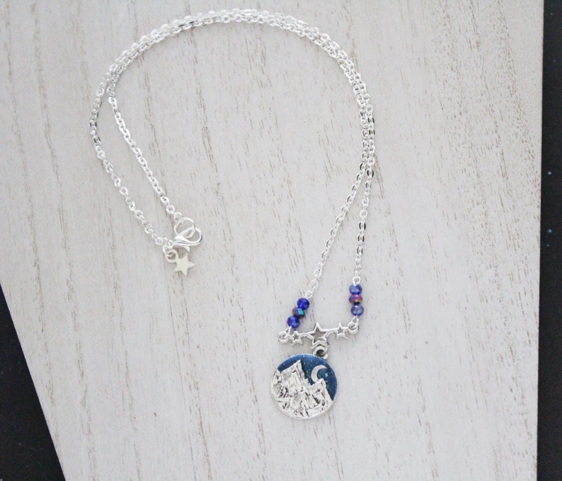 May include: A silver chain necklace with a silver charm featuring a mountain scene with a crescent moon and stars. The charm is surrounded by blue iridescent beads.
