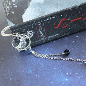 Puede incluir: Marcap&aacute;ginas de plata con un amuleto de cuervo, una cuenta negra y una cuenta roja colgando de una cadena. El marcap&aacute;ginas est&aacute; sobre un libro titulado "Six of Crows" de Leigh Bardugo.