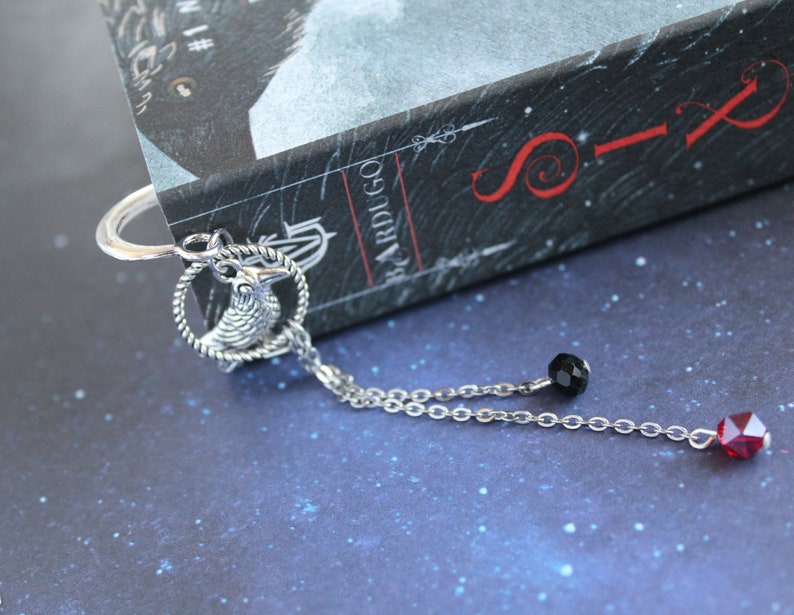 Puede incluir: Marcap&aacute;ginas de plata con un amuleto de cuervo, una cuenta negra y una cuenta roja colgando de una cadena. El marcap&aacute;ginas est&aacute; sobre un libro titulado "Six of Crows" de Leigh Bardugo.