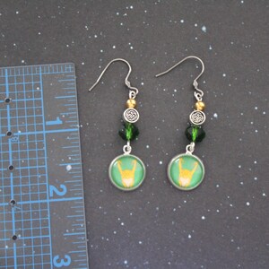 以下が含まれることがあります： シルバーカラーのフックが付いたぶら下がりピアス。各イヤリングは、緑色の背景に黄色の手のジェスチャーが付いた緑と金のデザインが特徴です。イヤリングには、緑と金のビーズとシルバーのケルトノットチャームが付いています。