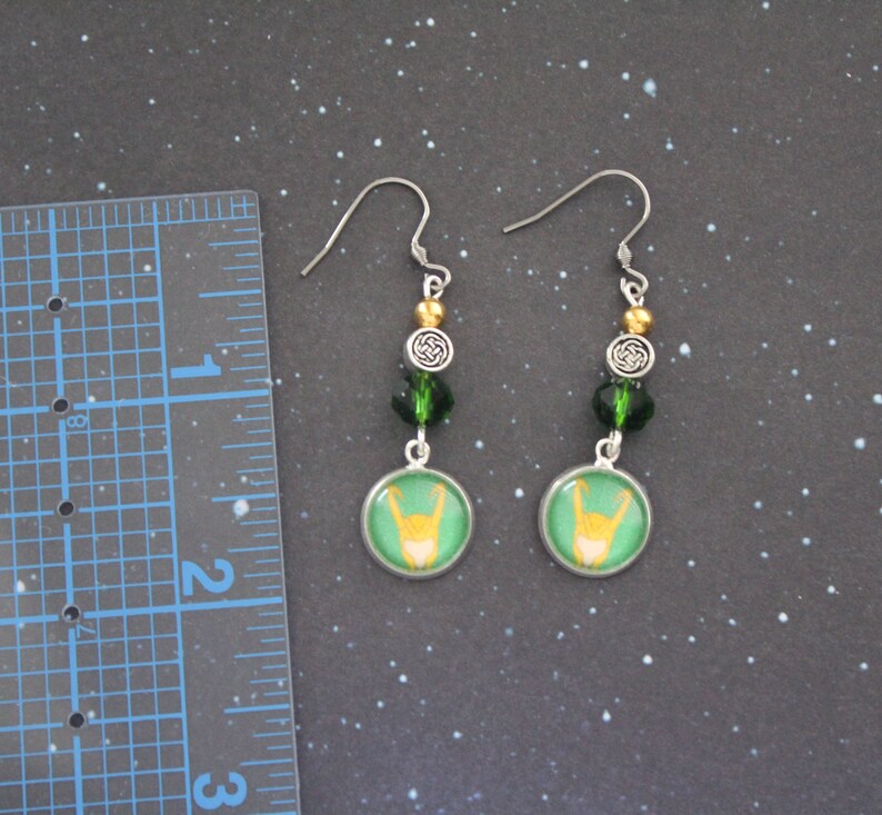 以下が含まれることがあります： シルバーカラーのフックが付いたぶら下がりピアス。各イヤリングは、緑色の背景に黄色の手のジェスチャーが付いた緑と金のデザインが特徴です。イヤリングには、緑と金のビーズとシルバーのケルトノットチャームが付いています。