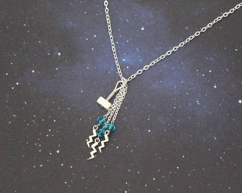 May include: A silver necklace with a silver hammer charm and blue crystal beads hanging from it. The necklace is on a dark blue background with white stars.