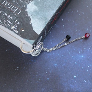 Puede incluir: Marcap&aacute;ginas de plata con un dise&ntilde;o decorativo y una cadena con cuentas rojas y negras. El marcap&aacute;ginas est&aacute; sujeto a un libro con una cubierta en blanco y negro.