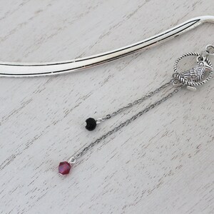 Puede incluir: Marcap&aacute;ginas de plata con una cadena colgante que presenta un peque&ntilde;o amuleto de p&aacute;jaro, una cuenta negra y una cuenta de cristal roja.