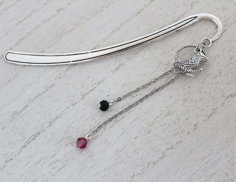 Puede incluir: Marcap&aacute;ginas de plata con una cadena colgante que presenta un peque&ntilde;o amuleto de p&aacute;jaro, una cuenta negra y una cuenta de cristal roja.