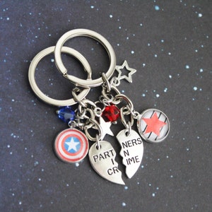 K&ouml;nnte beinhalten: Silberne Schl&uuml;sselanh&auml;nger mit Anh&auml;ngern. Zu den Anh&auml;ngern geh&ouml;ren ein Captain America-Schild, ein roter Stern und zwei halbherzf&ouml;rmige Anh&auml;nger mit der Aufschrift "PARTNERS IN CRIME". Zus&auml;tzliche Anh&auml;nger sind blaue und rote Perlen sowie sternf&ouml;rmige Anh&auml;nger.